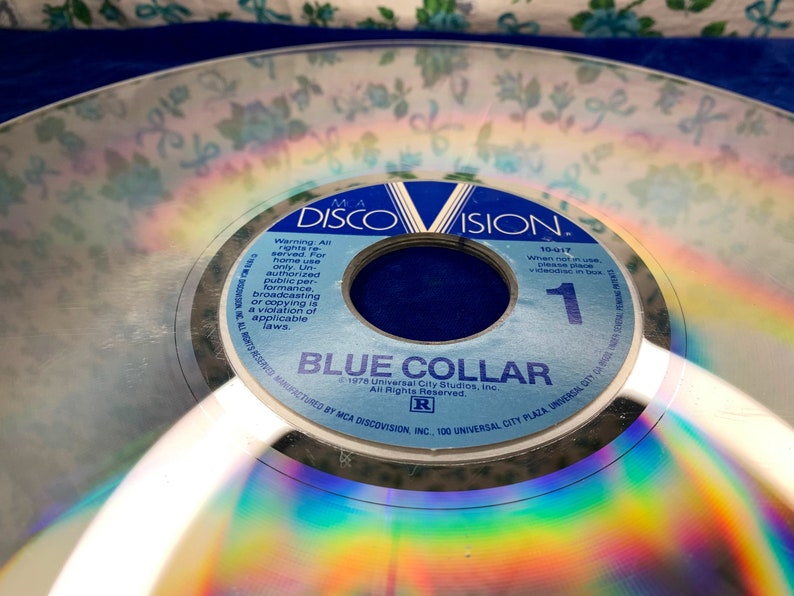 May include: A vintage DiscoVision disc with the title "Blue Collar." The disc has a reflective surface with rainbow hues. The label is blue and white, with the DiscoVision logo and copyright information.