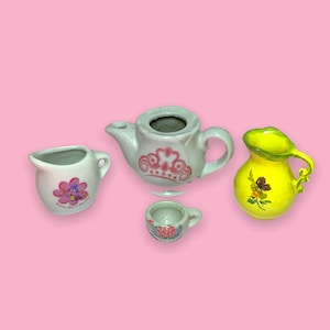 Puede incluir: Un juego de té de cerámica en miniatura sobre un fondo rosa. El juego incluye una tetera pequeña con un diseño de corona rosa, una crema con un gráfico de flores, una taza pequeña y una jarra amarilla con un diseño floral.