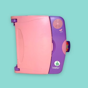 Puede incluir: Un juguete portátil de aprendizaje LeapPad rosa y morado con un cartucho insertado. El juguete tiene un altavoz, un botón de encendido/apagado y un asa. El logotipo de LeapFrog es visible en el lateral.
