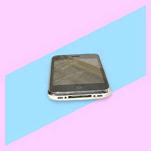 May include: A black smartphone with a cracked screen, resting on a blue and pink background. The phone has a silver trim and a visible home button. The screen is extensively damaged with multiple cracks.