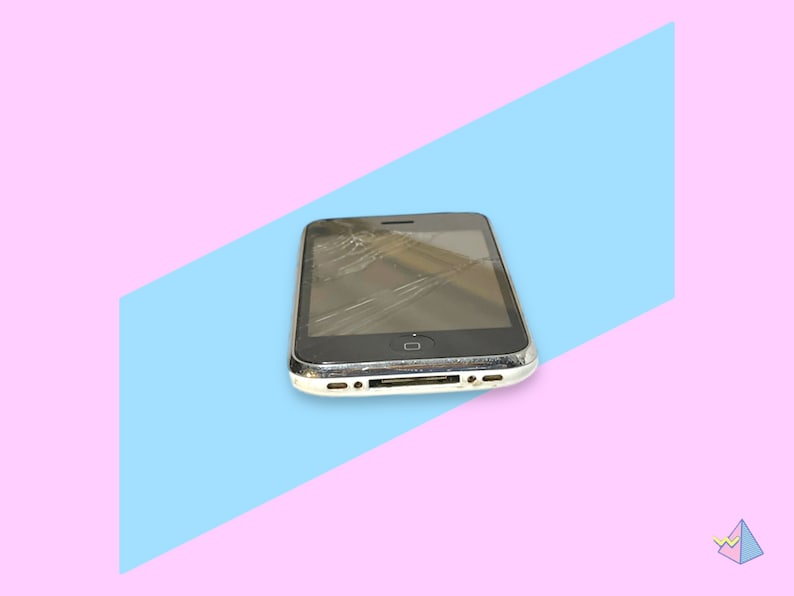 May include: A black smartphone with a cracked screen, resting on a blue and pink background. The phone has a silver trim and a visible home button. The screen is extensively damaged with multiple cracks.