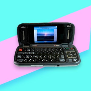 Peut inclure: Téléphone portable à clapet LG noir avec clavier coulissant, ouvert pour afficher une image de coucher de soleil. Le téléphone a un logo Verizon et un petit écran. Le fond est un motif géométrique turquoise et rose.