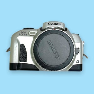 Pode incluir: C&acirc;mera Canon EOS IX Lite prateada com tampa de lente preta. O logotipo da Canon est&aacute; na parte superior e na tampa da lente. O nome do modelo "EOS IX Lite" est&aacute; impresso na lateral. Em um fundo azul claro.