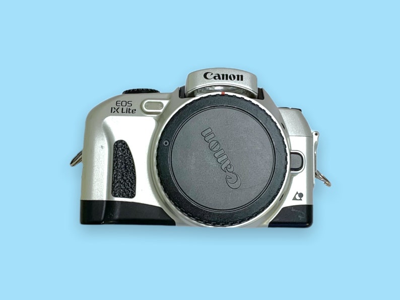 Pode incluir: C&acirc;mera Canon EOS IX Lite prateada com tampa de lente preta. O logotipo da Canon est&aacute; na parte superior e na tampa da lente. O nome do modelo "EOS IX Lite" est&aacute; impresso na lateral. Em um fundo azul claro.
