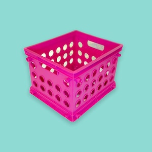 Puede incluir: Una caja de almacenamiento de plástico rosa brillante con numerosas perforaciones circulares. La caja tiene forma rectangular con un recorte de asa en cada extremo. El fondo es de color turquesa sólido.