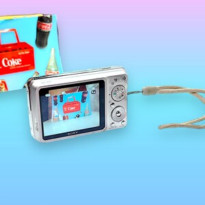May include: A silver digital camera with a beige wrist strap displays an image of a red Coke carrier with Coke bottles. A colorful background features a gradient of blue, pink, and purple.