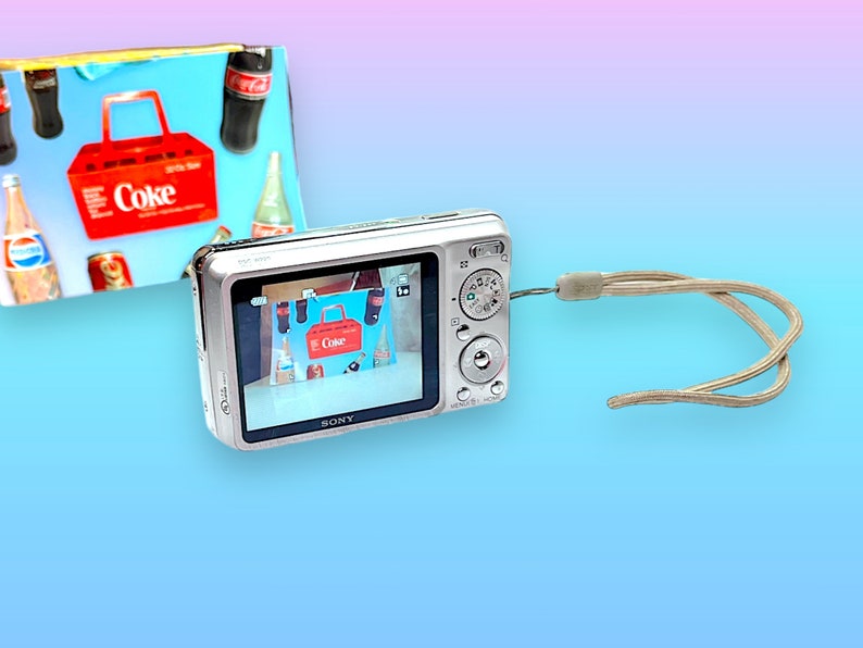 May include: A silver digital camera with a beige wrist strap displays an image of a red Coke carrier with Coke bottles. A colorful background features a gradient of blue, pink, and purple.