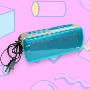 May include: A rectangular digital alarm clock with a translucent teal plastic casing and a black power cord. The top features small blue control buttons and a larger snooze button. The front display panel shows the word Advance in white text.