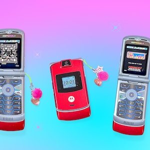 May include: Three retro Motorola flip phones in silver and red, with screens displaying video games. One red phone shows the time 12:21. Each phone has a charm with a pink pom-pom and heart. The background is a gradient of pink and blue.