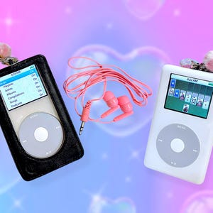 May include: Two digital music players with circular click wheels and screens displaying menus and card games, accompanied by a pair of pink wired earbuds. One player is encased in a black protective cover, while the other is white.