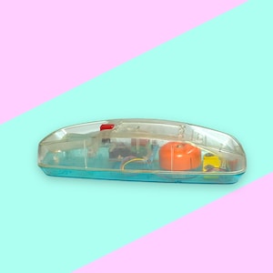 K&ouml;nnte beinhalten: Ein Vintage-Spielzeugtelefon aus klarem Kunststoff mit einem t&uuml;rkisfarbenen Sockel und einem roten Knopf. Die internen Komponenten des Telefons, einschlie&szlig;lich eines orangefarbenen Lautsprechers, sind durch die transparente Abdeckung sichtbar. Der Hintergrund ist diagonal in Rosa und T&uuml;rkis geteilt.