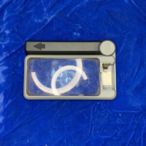 May include: A rectangular, gray handheld magnifier with a black handle and a clear lens. The lens has a white design in the center. The magnifier is on a blue surface.