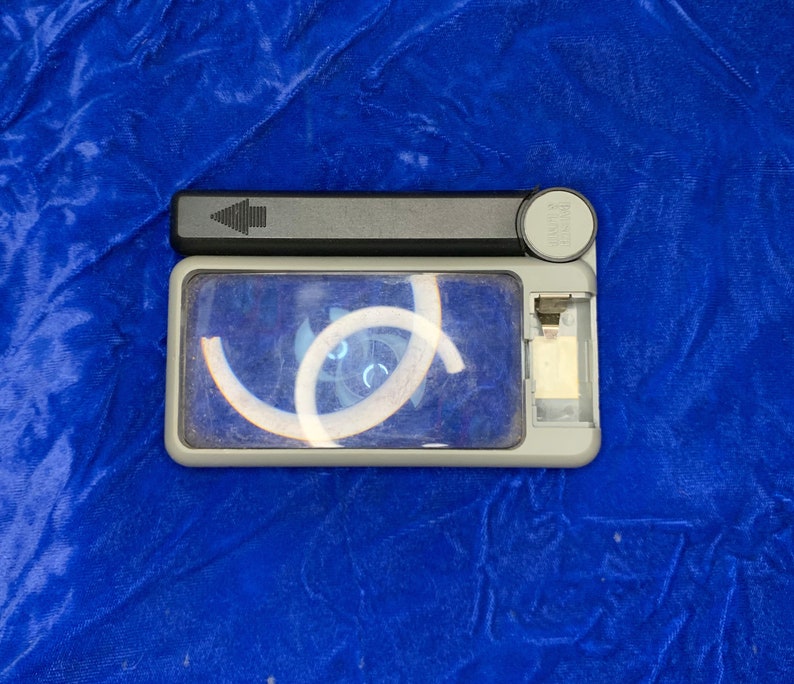 May include: A rectangular, gray handheld magnifier with a black handle and a clear lens. The lens has a white design in the center. The magnifier is on a blue surface.