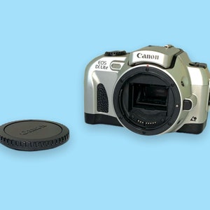 Pode incluir: C&acirc;mera Canon EOS IX Lite prateada com uma tampa de lente preta. O corpo da c&acirc;mera tem uma ader&ecirc;ncia texturizada e o logotipo da Canon &eacute; vis&iacute;vel. A tampa da lente &eacute; preta com o logotipo da Canon.