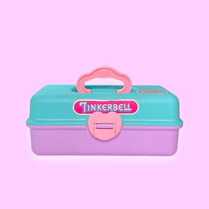 Puede incluir: Una caja de herramientas Tinkerbell de plástico de dos tonos, en verde azulado y lavanda, con un asa y cierre rosas. La caja de herramientas presenta el logotipo de Tinkerbell en un óvalo rosa y blanco. El fondo es rosa claro liso.