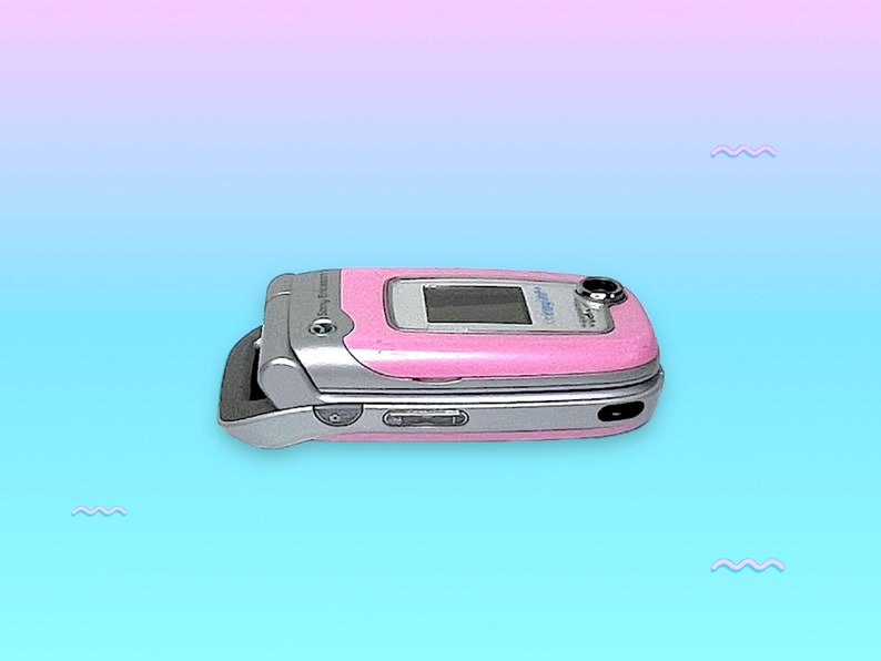 May include: A pink and silver clamshell cell phone. The phone is open, revealing a small screen. The phone has a silver hinge and a pink exterior. The phone is a vintage model.