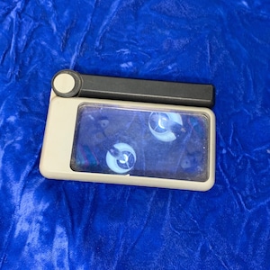 May include: A rectangular, off-white magnifying glass with a black handle and a circular button. The glass area shows a distorted reflection. The item is set against a blue, textured background.