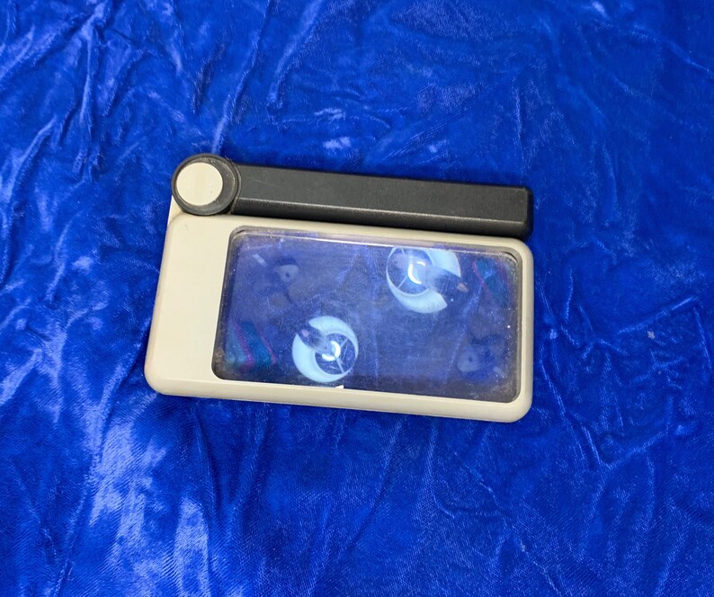 May include: A rectangular, off-white magnifying glass with a black handle and a circular button. The glass area shows a distorted reflection. The item is set against a blue, textured background.