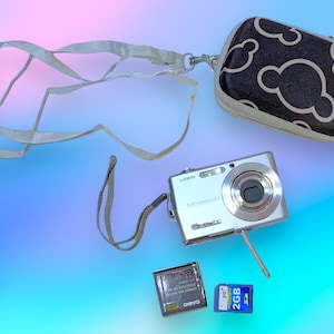 Może przedstawiać: Srebrny aparat cyfrowy Casio z matrycą 5,0 megapikseli, pasującym etui z wzorem chmur, kartą SD 2 GB i baterią. Aparat posiada pasek na nadgarstek i pasek na szyję.
