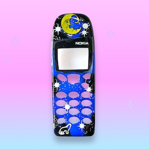 May include: A black Nokia phone with a custom painted design. The design features a blue night sky with stars, a crescent moon with a face, and a howling wolf. The phone has a blank screen and pink buttons.