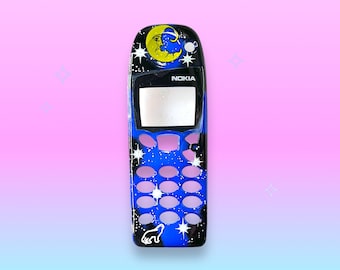 May include: A black Nokia phone with a custom painted design. The design features a blue night sky with stars, a crescent moon with a face, and a howling wolf. The phone has a blank screen and pink buttons.