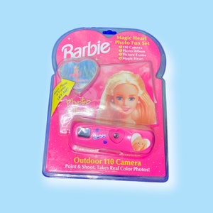Puede incluir: Un conjunto fotográfico Barbie Magic Heart rosa, que incluye una cámara 110, un álbum de fotos y un marco de fotos. El embalaje presenta una imagen de muñeca Barbie y el texto "Outdoor 110 Camera, Point & Shoot, Takes Real Color Photos!"