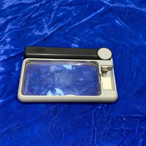 May include: A rectangular magnifying glass with a gray frame, black handle, and a small white button. The lens is clear, and the handle has a small, textured area. The magnifying glass is on a blue surface.
