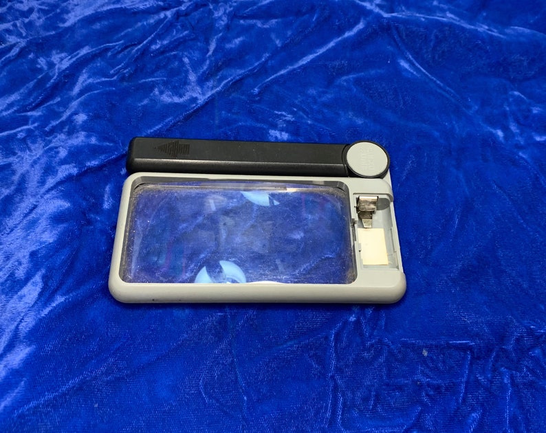 May include: A rectangular magnifying glass with a gray frame, black handle, and a small white button. The lens is clear, and the handle has a small, textured area. The magnifying glass is on a blue surface.