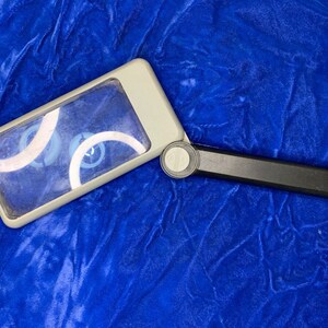 May include: A rectangular magnifying glass with a gray frame and a black handle. The lens is clear, and the handle folds for storage. The magnifying glass is set against a blue background.