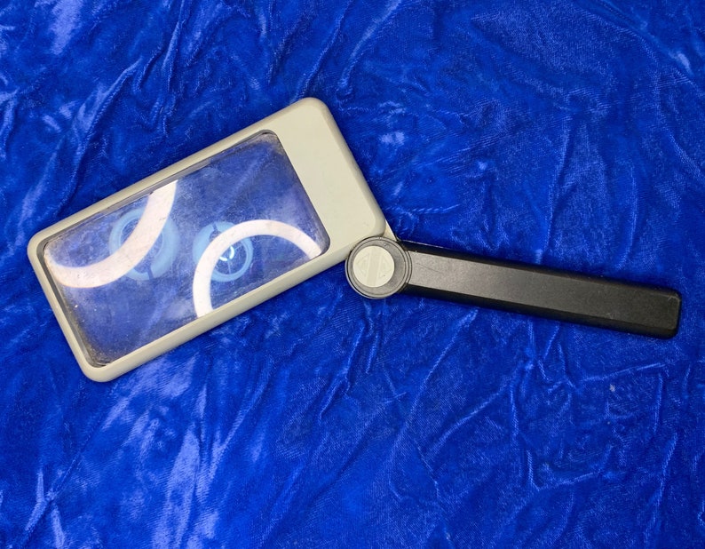 May include: A rectangular magnifying glass with a gray frame and a black handle. The lens is clear, and the handle folds for storage. The magnifying glass is set against a blue background.