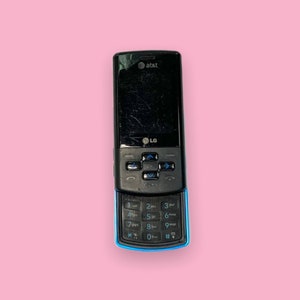 May include: A black LG flip phone with a blue accent on a pink background. The phone has a keypad and a screen with the AT&T logo. The phone shows signs of wear and tear.