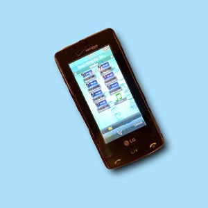 Peut inclure: Un téléphone portable LG C710 marron foncé avec un logo Verizon en haut. L'écran affiche un menu avec des icônes et le texte "Searching for Svc... Media". Le téléphone est sur un fond bleu clair.