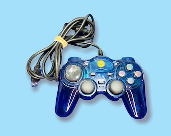 May include: A transparent blue video game controller with a black cable and a USB connector. The controller has multiple buttons, including a clear button, and two grey analog sticks. A yellow sticker is visible on the controller.