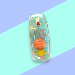 K&ouml;nnte beinhalten: Ein Vintage-Spielzeugtelefon aus klarem Kunststoff mit einem orangefarbenen Zifferblatt und einer roten Taste. Die internen Komponenten des Telefons sind sichtbar. Der Hintergrund ist hellblau und lila geteilt.