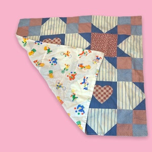 Puede incluir: Un edredón patchwork en tonos azules, rojos y blancos. Presenta patrones de estrellas y cuadrados, con un parche en forma de corazón. La parte inferior es blanca con ilustraciones coloridas con temática de circo.