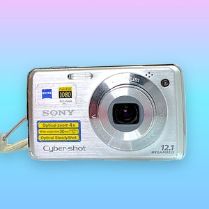 May include: Silver Sony Cyber-shot digital camera with a Zeiss lens. Features include 12.1 megapixels, optical zoom 4x, and a wide-angle lens of 30mm. Includes a wrist strap and the words "Full HD 1080" on the top left.