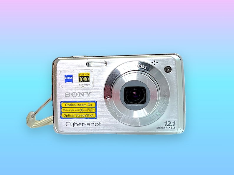 May include: Silver Sony Cyber-shot digital camera with a Zeiss lens. Features include 12.1 megapixels, optical zoom 4x, and a wide-angle lens of 30mm. Includes a wrist strap and the words "Full HD 1080" on the top left.