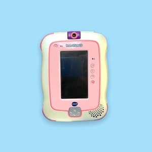 Può includere: Un tablet VTech InnoTab rosa e crema per bambini, con schermo nero, fotocamera viola e pulsanti funzione. Il tablet ha un design arrotondato e un altoparlante in basso a destra.