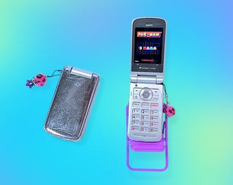 May include: Two silver flip phones, one open displaying a Pac-Man game, the other closed. Both phones have keychains with pink and black pom-poms. One phone is propped on a small purple chair. The background is a gradient of blue and green.