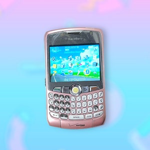 以下が含まれることがあります： ローズゴールドのBlackBerryスマートフォン。フルQWERTYキーボードとアイコンを表示する画面があります。Verizonのロゴと中央のナビゲーションボタンが付いています。画面には7:19と表示されています。