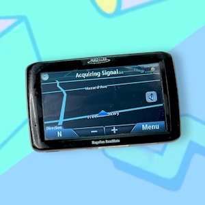 Puede incluir: Un dispositivo GPS Magellan RoadMate negro que muestra un mapa con el texto "Acquiring Signal". La pantalla muestra nombres de calles y un botón de menú. El dispositivo está sobre un fondo colorido.