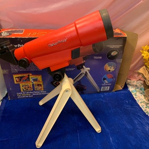 May include: A vintage Fisher-Price toy telescope set. The red telescope is mounted on a cream-colored tripod. The box behind the telescope has the words "Fisher-Price" and "Fun Starters" printed on it.