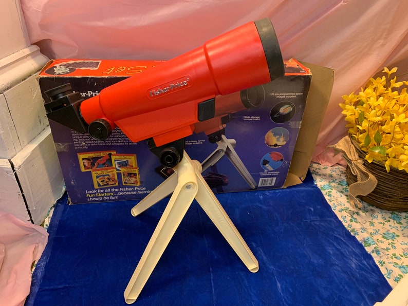 May include: A vintage Fisher-Price toy telescope set. The red telescope is mounted on a cream-colored tripod. The box behind the telescope has the words "Fisher-Price" and "Fun Starters" printed on it.