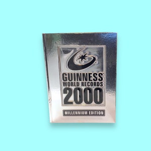 Peut inclure: Un livre Guinness World Records 2000 Édition du Millénaire encadré d'argent. La couverture présente le logo Guinness avec une étoile et un design incurvé. Le texte "GUINNESS WORLD RECORDS 2000" est bien en évidence, avec "MILLÉNNIUM EDITION" en dessous.