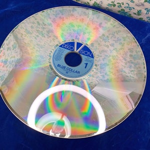 May include: A vintage Discovision disc with a floral pattern overlay. The disc has a blue and white label with the text "Blue Collar" and the number "1". The disc reflects a rainbow of colors.