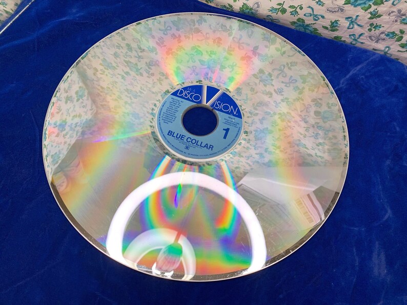 May include: A vintage Discovision disc with a floral pattern overlay. The disc has a blue and white label with the text "Blue Collar" and the number "1". The disc reflects a rainbow of colors.