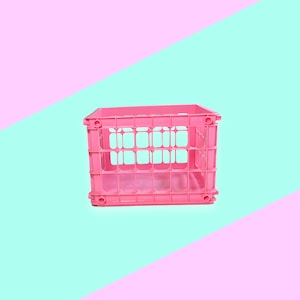 Puede incluir: Una caja de plástico rosa brillante con forma rectangular y lados abiertos. La caja se encuentra sobre un fondo de colores pastel rosa y turquesa en diagonal, creando una estética vibrante y juguetona.