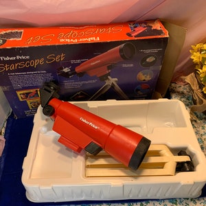 May include: A red Fisher-Price Starscope Set, designed for kids, is displayed in its original box and packaging. The telescope is red with black accents and sits in a white plastic tray. The box features images of space and the words "Starscope Set".