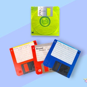 Puede incluir: Vista cenital de cuatro disquetes de 3,5 pulgadas vintage. Uno es verde neón, los otros son rojos, azules y grises. Cada disco tiene una etiqueta con texto manuscrito. El disco verde tiene el logotipo de Imation.
