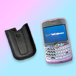 Peut inclure: Un smartphone BlackBerry rose avec un clavier complet QWERTY et un écran affichant le logo Verizon Wireless. À côté, un étui en cuir noir avec le logo BlackBerry.